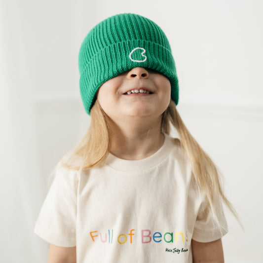 Child wearing a green beanie and a cream t-shirt with colourful text that says 'full of beans' on a plain background