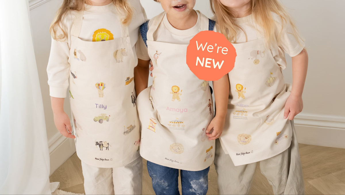Three young children standing indoors wearing matching cream-colored pajama sets with small yellow and blue printed motifs. The center child holds a red circular sticker that reads 'We're NEW'.
