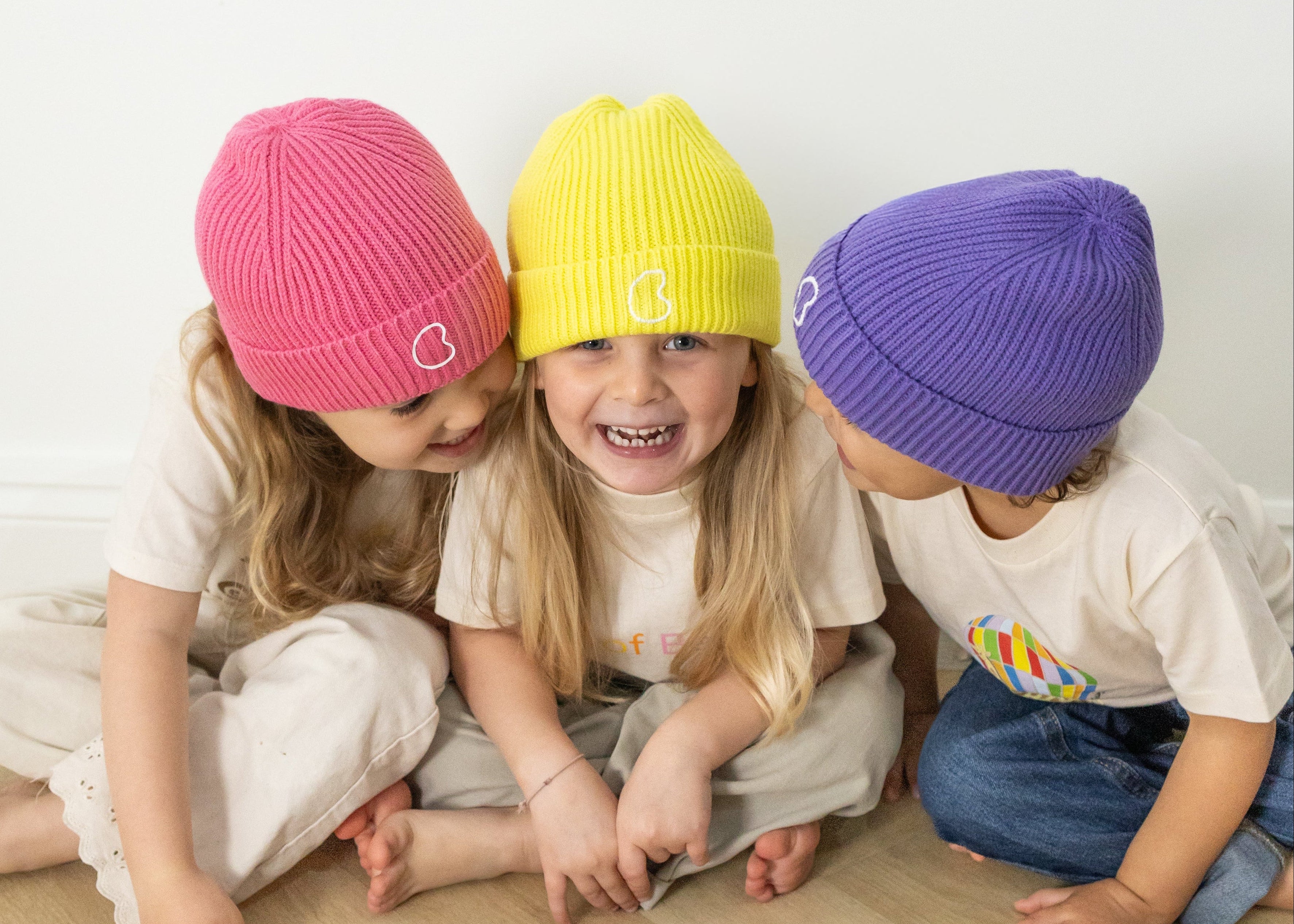 Three children wearing colourful beanies sitting on a wooden floor.