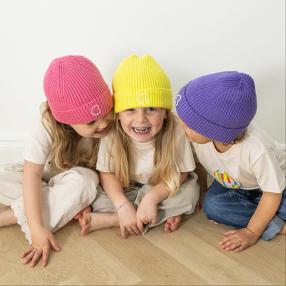 Three children wearing colourful beanies sitting on a wooden floor.