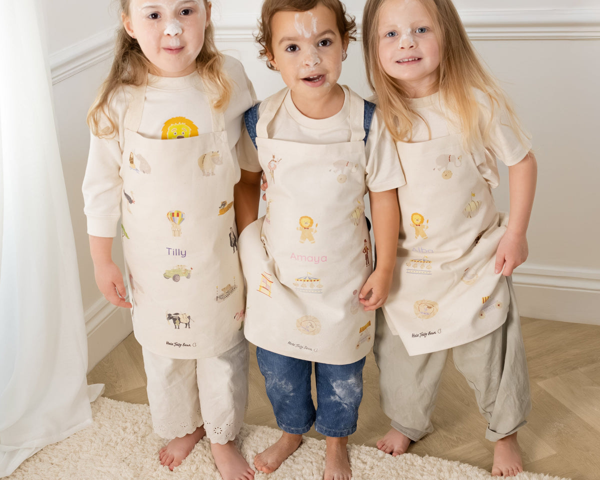 Three young children standing barefoot indoors on a cream rug, each wearing a matching beige apron with small printed motifs over light-colored outfits.