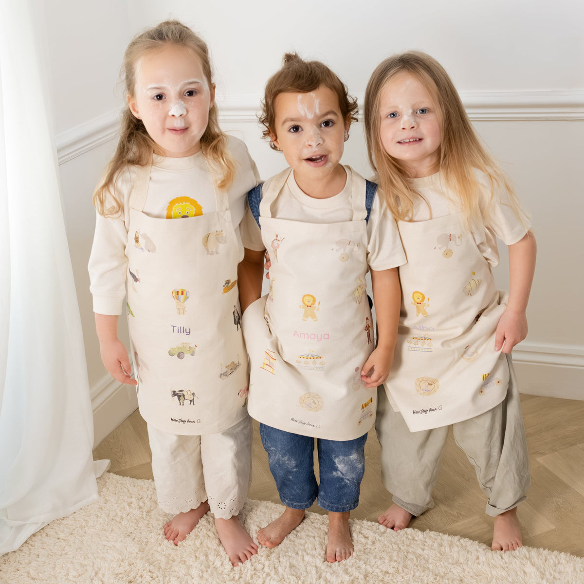 Three young children standing barefoot indoors on a cream rug, each wearing a matching beige apron with small printed motifs over light-colored outfits.
