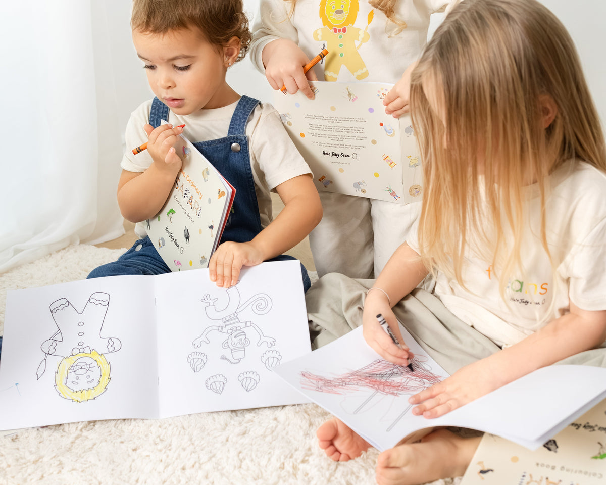 Three children sitting on a white carpet, coloring in picture books with crayons. The books feature colorful illustrations of animals and characters, and the children are wearing matching white t-shirts with Hele Jelly Bean branding.