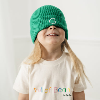 Child wearing a green beanie and a cream  t-shirt with colourful text that says 'full of beans' on a plain background