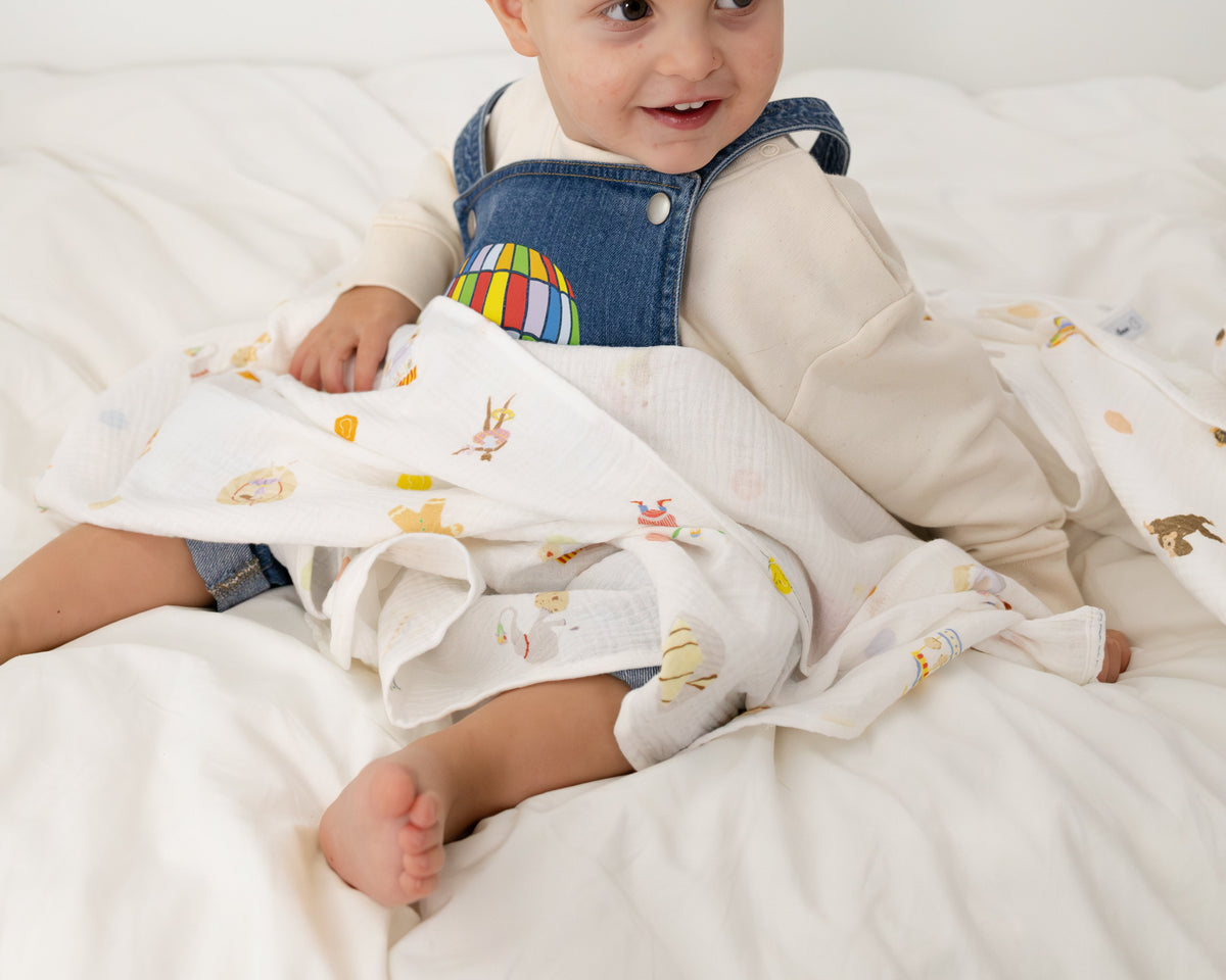 A baby sitting on white bedding, wrapped in a white muslin blanket with a colorful cartoon animal print. The baby is wearing a cream long-sleeve shirt and denim overalls.