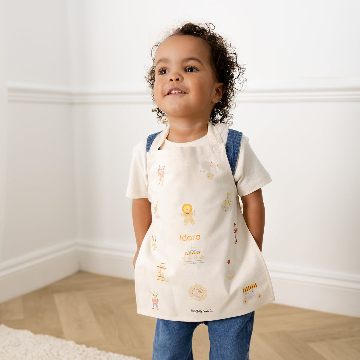 A toddler stands barefoot on a white shag rug in a bright room, wearing a cream bib-style tunic with small multicolor illustrations, blue denim pants, and blue shoulder straps.