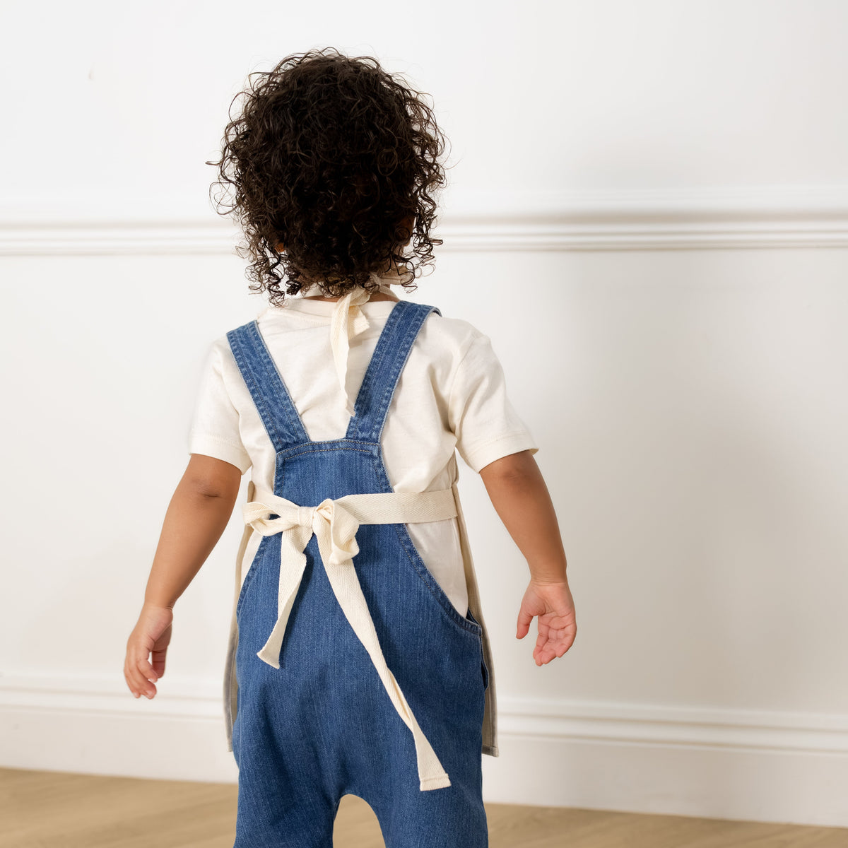 A toddler viewed from behind wearing a white short-sleeve top and blue denim-style overalls with a cream tie belt, standing barefoot on a light wood floor in a bright room.